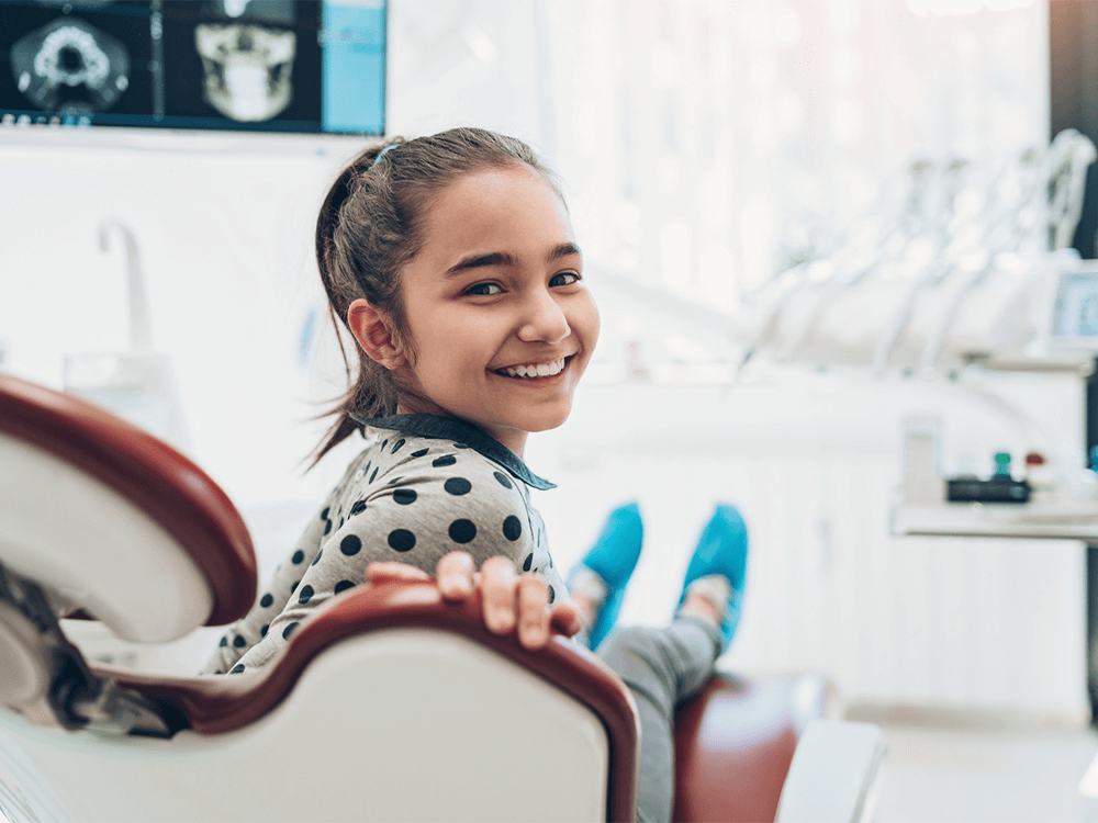 A young girl sitting in a dental chair, smiling at the camera. She is wearing a polka dot shirt and has her hair tied back in a ponytail. The background shows dental equipment and a computer monitor displaying dental X-rays, creating a professional and clean dental office setting. The girl appears relaxed and happy, indicating a positive experience at the dentist.