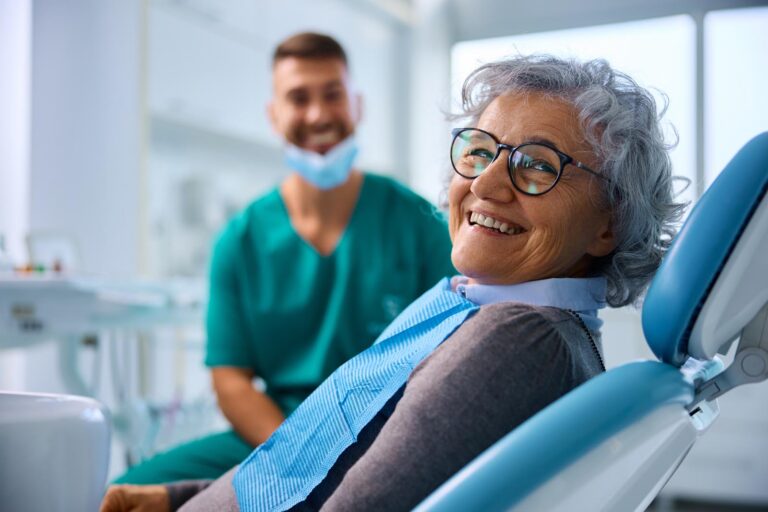 Older patient in dental chair wearing glasses and blue bib, with dental professional in green scrubs visible in background clinic setting.