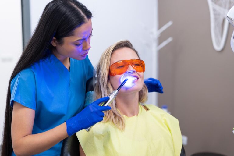 Dental professional in blue scrubs using curing light on patient wearing orange protective glasses and yellow bib in clinic setting.